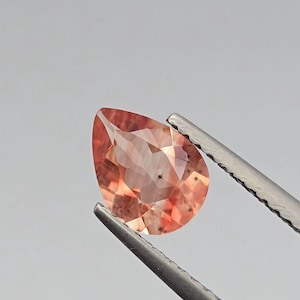 May include: A faceted, pear-shaped gemstone with a peach and orange color is held by tweezers against a gray background. The gem has internal inclusions and a polished surface, reflecting light.