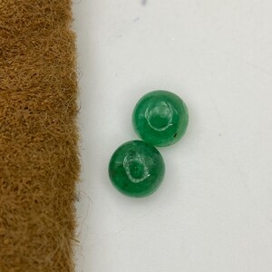 May include: Two round, polished emerald gemstones. The gems are a vibrant green color and appear to be cabochon cut. The background is a plain white surface, with a brown, textured material in the upper left corner.