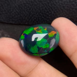 May include: A dark, oval-shaped opal gemstone with a polished surface. The opal exhibits flashes of green, orange, and blue. The gemstone is held in a hand, highlighting its colour and shape. The opal is a beautiful example of its type.