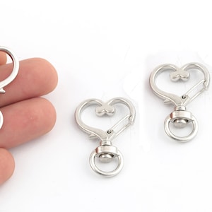 May include: Three silver heart-shaped swivel snap hooks with a ring on the bottom. The hooks are designed to be used for attaching keys, bags, or other items.