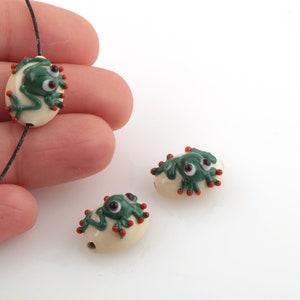 Puede incluir: Tres cuentas de vidrio con ranas verdes sobre un fondo blanco. Las ranas tienen manchas rojas y ojos blancos.