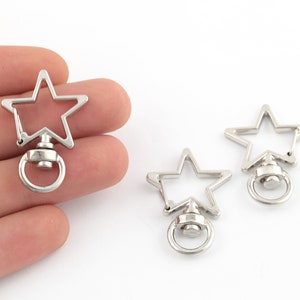 May include: Two silver star-shaped swivel snap hooks with a ring on the bottom. The hooks are open and ready to be attached to a keychain, bag, or other item.