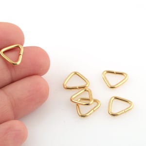 May include: A close-up of six gold-toned triangle-shaped metal rings. The rings are small and have a smooth, shiny finish.