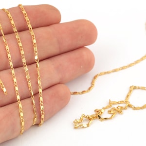 May include: A gold chain necklace with a delicate, flat, oval-shaped link design. The chain is shown laid out on a hand and on a white surface.