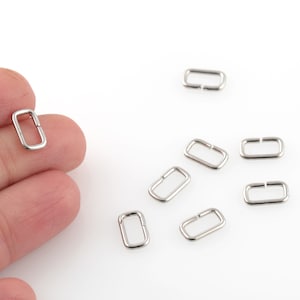 Puede incluir: Primer plano de diez anillos de salto rectangulares de metal plateado. Los anillos miden aproximadamente 1,2 cm de largo y 0,6 cm de ancho.