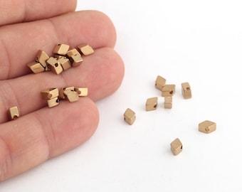 May include: A pile of gold-colored metal beads with a hole in the center of each bead. The beads are in the shape of a diamond. The beads are scattered on a white surface.