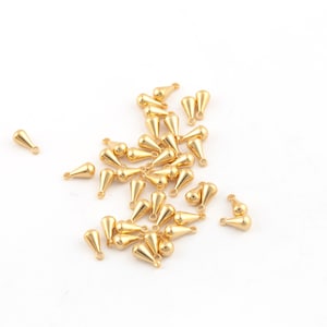 May include: A group of gold teardrop-shaped metal charms with a loop at the top. The charms are scattered on a white surface.