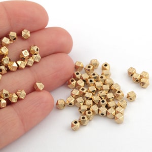 May include: A pile of gold-colored metal beads with a faceted, geometric design. The beads have a hole in the center for stringing.