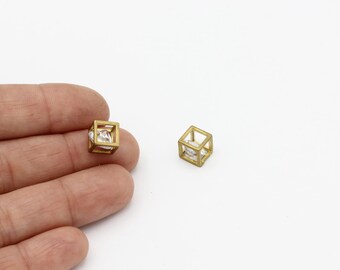 Puede incluir: Un par de pendientes de tono dorado con una piedra de circonita cúbica transparente dentro de una forma de cubo geométrico.
