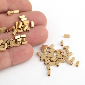 Könnte beinhalten: Ein Haufen goldfarbener Metall-Spacer-Perlen, einige befinden sich in einer Hand, andere sind auf einer weißen Oberfläche verstreut.