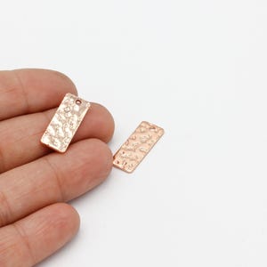 May include: Two rose gold-colored metal rectangular tags with a hammered texture. The tags have a hole at the top for attaching to a necklace or bracelet.