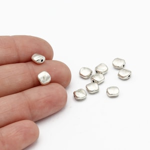 May include: A group of silver-toned metal beads with a slightly irregular, rounded square shape. The beads have a hole in the center for stringing.