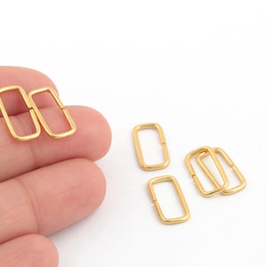 May include: Gold-toned metal rectangular rings, also known as D-rings, are shown on a white background. The rings are approximately 1 inch long and 1/2 inch wide.