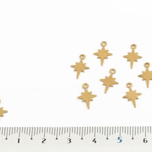 May include: Eight gold-toned metal star charms with a pointed tip, arranged in a circle on a white surface. A ruler is placed below the charms, with the numbers 0 through 7 visible.