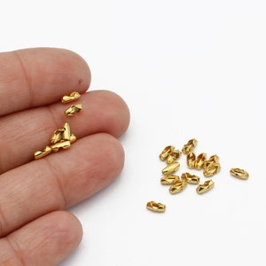 May include: Small, gold-colored metal chain connectors scattered on a white surface. The connectors have a geometric shape with multiple facets. Some are held in a hand, while others are loose.
