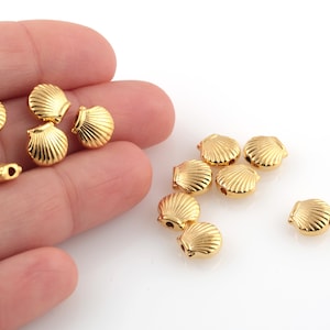 May include: A group of gold-colored seashell-shaped beads with a hole in the center. The beads are scattered on a white surface.