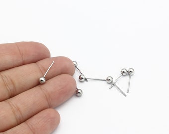 Puede incluir: Primer plano de una mano sosteniendo un pendiente de bola de plata. El pendiente es pequeño y tiene un diseño sencillo. Hay varios pendientes más esparcidos sobre una superficie blanca.