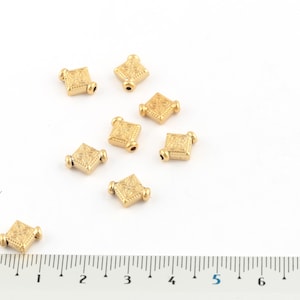 May include: Eight gold-toned square beads with intricate designs. The beads are approximately 1 cm in size and have a hole in the center for stringing.