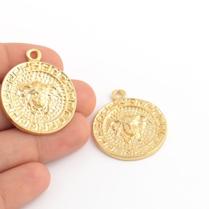 May include: Two gold-tone metal pendants with a raised design of a face with a crown. The pendants are round and have a detailed border.