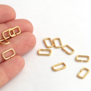 May include: A group of gold-colored metal rectangular rings, also known as D-rings, are scattered on a white surface. The rings are about 1 inch long and 1/2 inch wide.