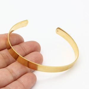 May include: A gold-colored metal bangle bracelet with a smooth, flat surface. The bracelet is open on one side and has two small holes on the ends.
