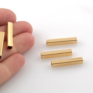 Pode incluir: Quatro tubos metálicos dourados, cada um com uma seção transversal quadrada. Os tubos medem aproximadamente 2,5 cm de comprimento e estão dispostos em um padrão escalonado.
