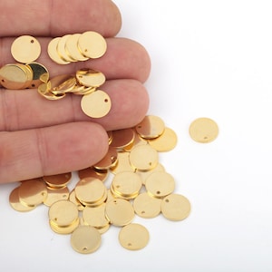 May include: A pile of gold-colored metal discs with a small hole in each one. The discs are scattered on a white surface.