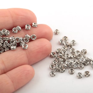 May include: A close-up of a pile of silver-toned metal beads with clear rhinestones. The beads are round and have a hole in the center. They are scattered on a white surface.