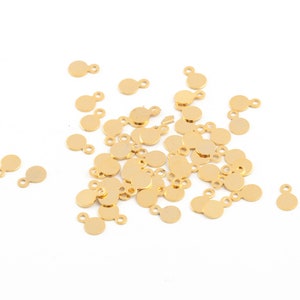 Pode incluir: Um grande grupo de pingentes circulares de metal dourado com um pequeno laço de cada lado. Os pingentes estão espalhados sobre uma superfície branca.