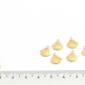 May include: Seven gold-colored metal seashell charms with a loop at the top for attaching to jewelry or other crafts. The charms are arranged on a white surface with a ruler for scale.