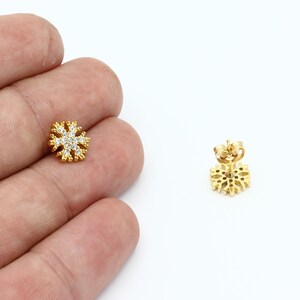 May include: A pair of gold snowflake earrings with clear cubic zirconia stones. The earrings are shown on a white background.