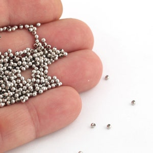 May include: A hand holding a large quantity of small, silver metal beads. Some beads are scattered on a white surface.