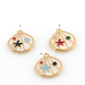 Puede incluir: Tres abalorios de metal de color dorado con forma de conchas marinas con esmalte blanco y detalles de esmalte de colores. Cada abalorio presenta una estrella de mar de color diferente y un punto de color diferente.