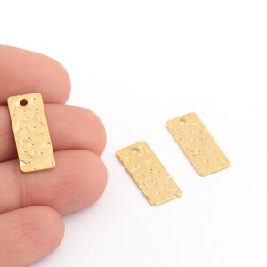 May include: Three gold-colored rectangular charms with a textured surface and a hole at the top. One charm is held in a hand, while the other two are on a white background. These charms are suitable for jewelry making.