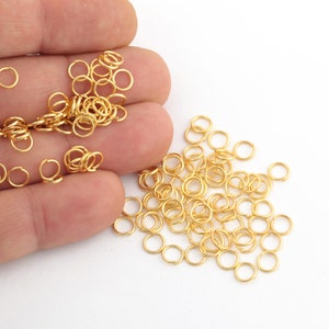 May include: A large pile of gold jump rings, a type of jewelry finding, on a white surface. The jump rings are small and round, and they are perfect for connecting different pieces of jewelry together.