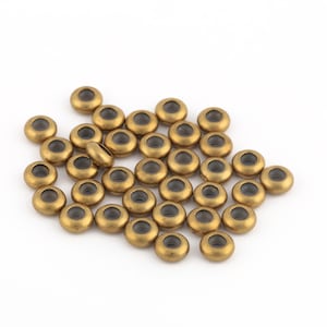 Puede incluir: Un montón de cuentas de metal de color dorado con un agujero central transparente. Las cuentas son redondas y tienen un acabado liso y brillante.