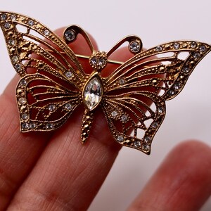May include: A gold-toned butterfly brooch with intricate openwork wings. The brooch is embellished with numerous small, clear crystals and a larger, teardrop-shaped crystal at the center. The butterfly's antennae are also adorned with crystals.
