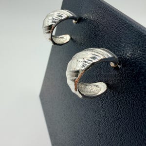 Puede incluir: Un par de pendientes de aro plateados. Cada pendiente tiene un diseño texturizado y acanalado en la superficie exterior. Los pendientes se muestran sobre una superficie negra sobre un fondo blanco.