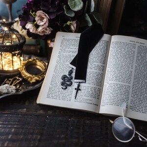 May include: A black velvet ribbon bookmark featuring a silver-toned snake charm with small star accents and a small silver-toned cross charm dangling from the bottom edge, resting on the open pages of a printed book with visible text.