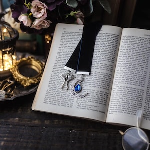 May include: Two black velvet ribbon bookmarks resting on an open book. One bookmark features a silver moth charm, while the other displays a silver crescent moon charm with a dangling blue teardrop-shaped gemstone accent at the center.