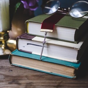 K&ouml;nnte beinhalten: Ein Stapel von drei B&uuml;chern mit einem roten Lesezeichen und einem silbernen Schwertlesezeichen. Das Lesezeichen h&auml;ngt &uuml;ber den Rand des obersten Buches. Die B&uuml;cher liegen auf einem Holztisch.
