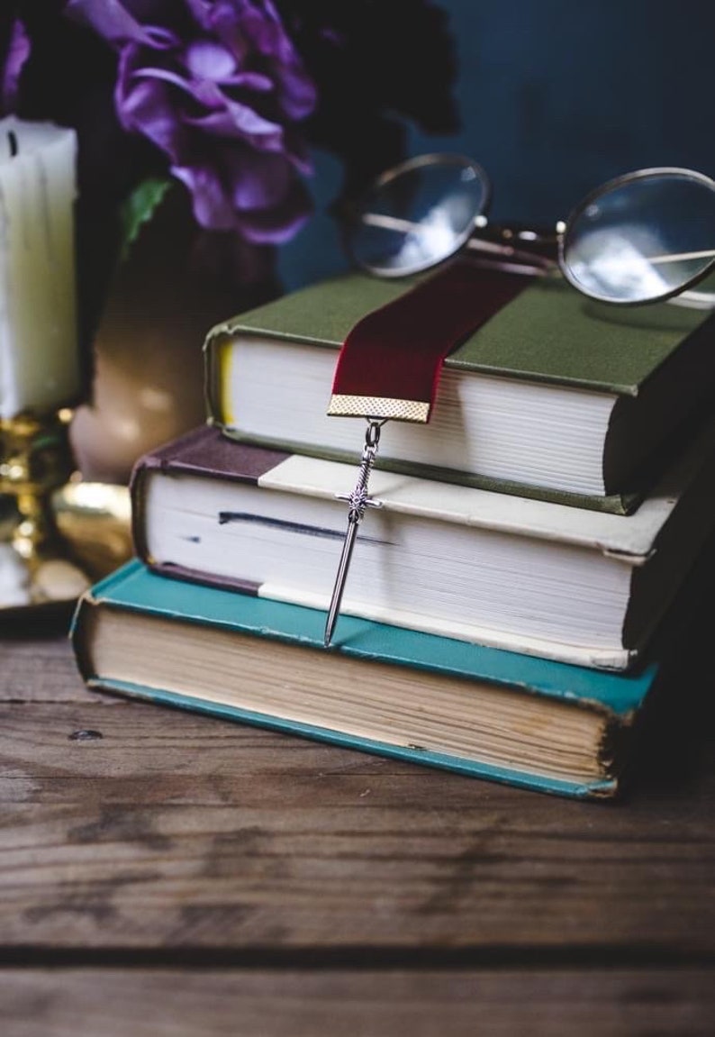 K&ouml;nnte beinhalten: Ein Stapel von drei B&uuml;chern mit einem roten Lesezeichen und einem silbernen Schwertlesezeichen. Das Lesezeichen h&auml;ngt &uuml;ber den Rand des obersten Buches. Die B&uuml;cher liegen auf einem Holztisch.