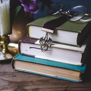 K&ouml;nnte beinhalten: Ein Stapel von drei alten B&uuml;chern mit einem burgunderfarbenen Samtlesezeichen und einem Drachenanh&auml;nger. Das oberste Buch ist gr&uuml;n, das mittlere lila und das untere t&uuml;rkis. Eine runde Brille liegt oben auf.
