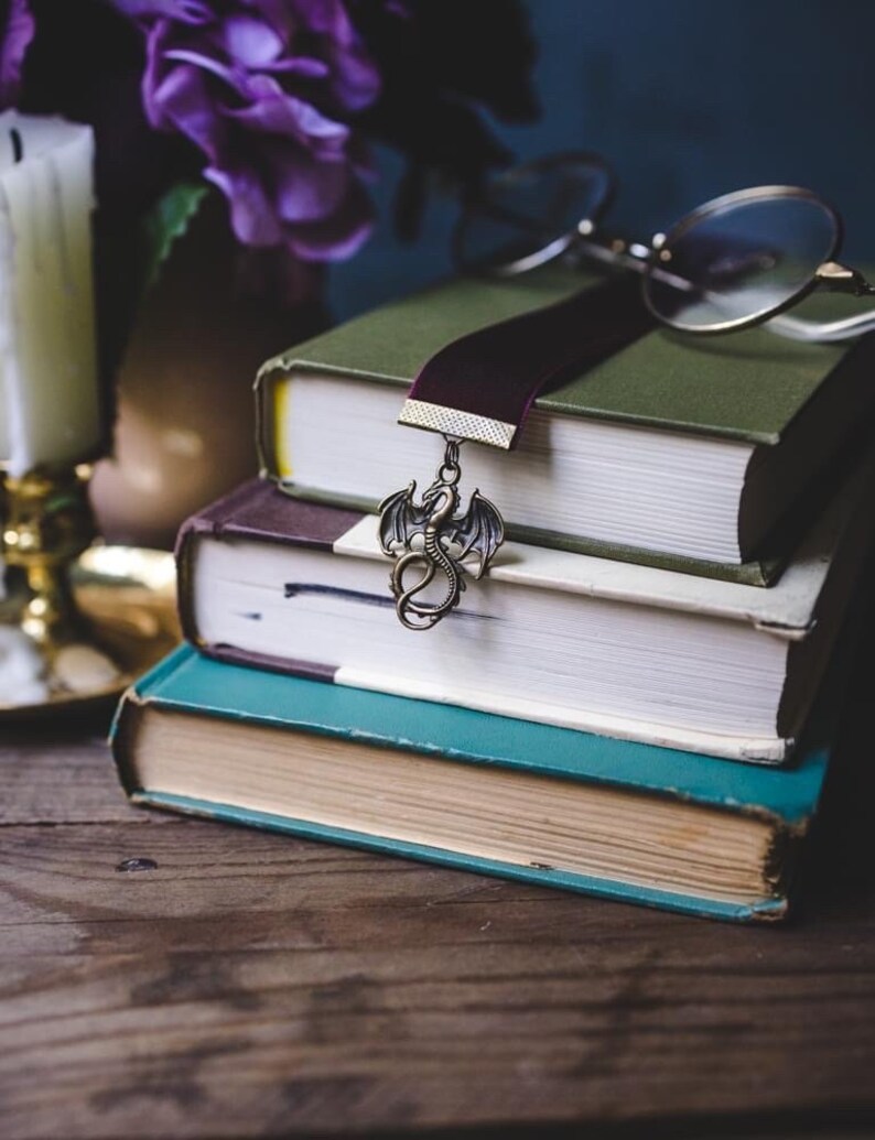 K&ouml;nnte beinhalten: Ein Stapel von drei alten B&uuml;chern mit einem burgunderfarbenen Samtlesezeichen und einem Drachenanh&auml;nger. Das oberste Buch ist gr&uuml;n, das mittlere lila und das untere t&uuml;rkis. Eine runde Brille liegt oben auf.
