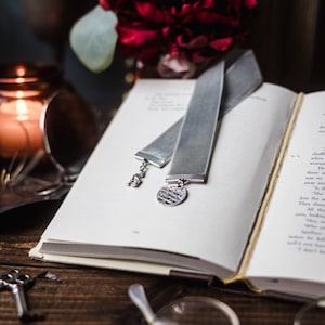May include: A grey velvet bookmark with a silver charm and a silver inscription. The bookmark is lying on an open book with a red flower in the background.