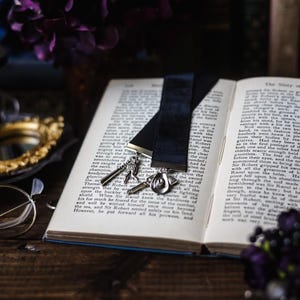 May include: An open book with a black velvet bookmark and a silver charm attached. The book is on a wooden table with a purple flower and a pair of glasses.