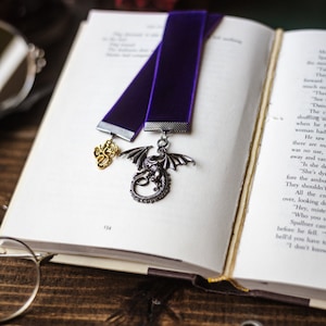 May include: A purple velvet bookmark with a silver dragon and a gold scorpion charm. The bookmark is laying on an open book with a pair of glasses and a key on a wooden surface.