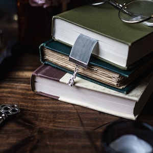 May include: A stack of three books with a silver bookmark and a pair of glasses on top. The books are green, blue, and brown. The glasses are round and have a gold frame.