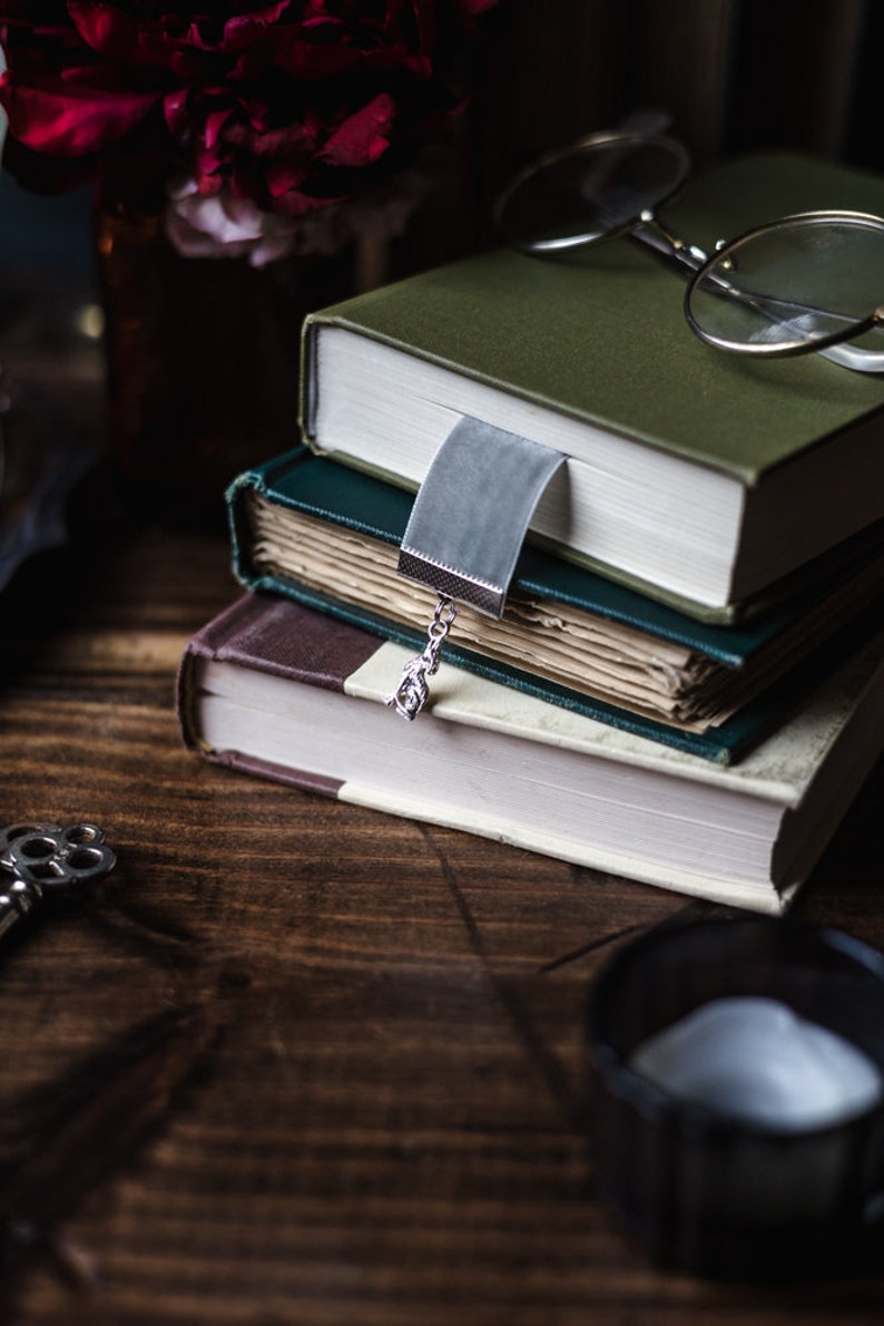 May include: A stack of three books with a silver bookmark and a pair of glasses on top. The books are green, blue, and brown. The glasses are round and have a gold frame.