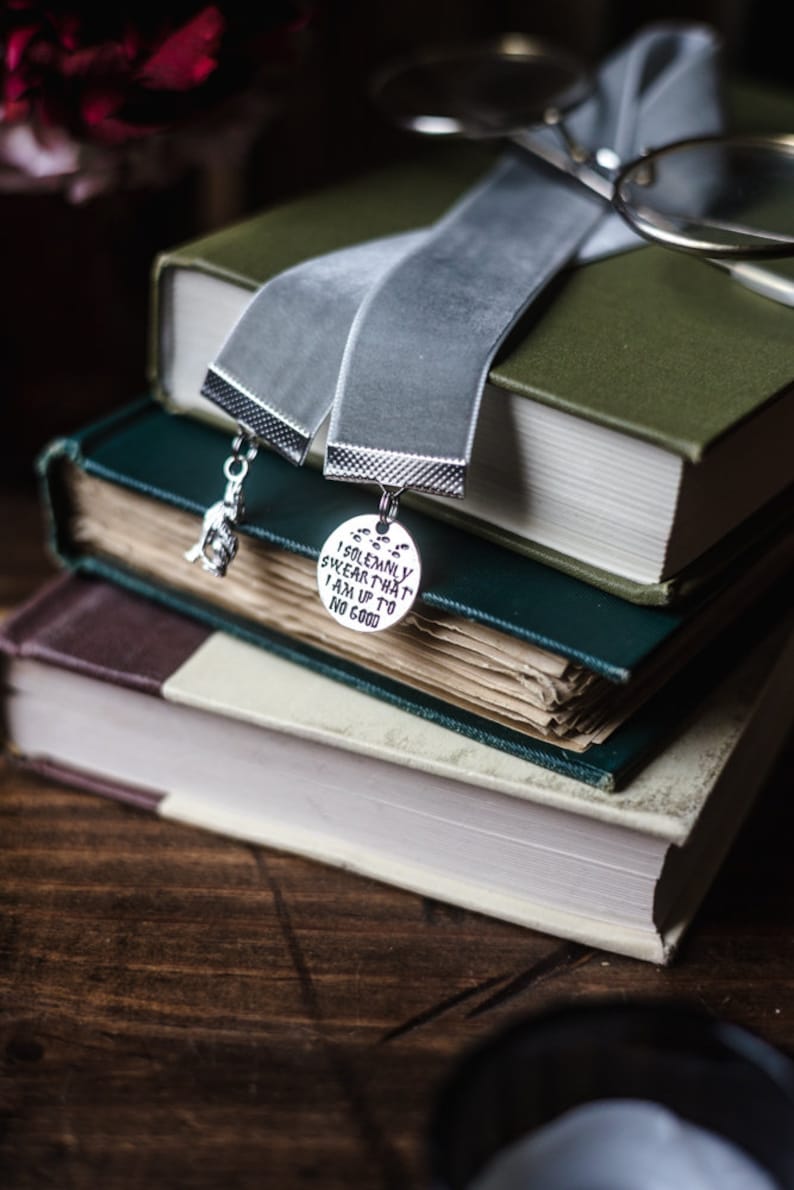 May include: A stack of three books with a grey velvet bookmark. The bookmark has two charms, one with the text "I solemnly swear that I am up to no good" and the other with a small figure.